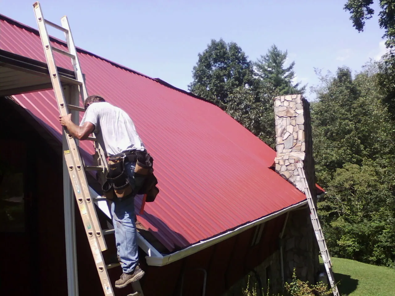 Roofer on a ladder beside a red metal roof and stone chimney — Soffit & Fascia Repair by Roofing Brothers in Scio